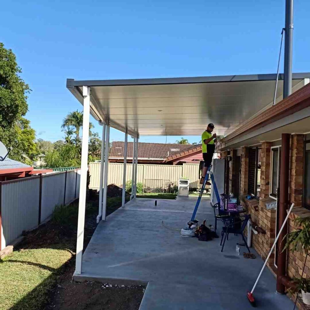 A person in a neon shirt stands on a ladder installing a white roof over a concrete patio attached to a brick house. There are tools and a broom nearby, with a backyard and trees in the background.